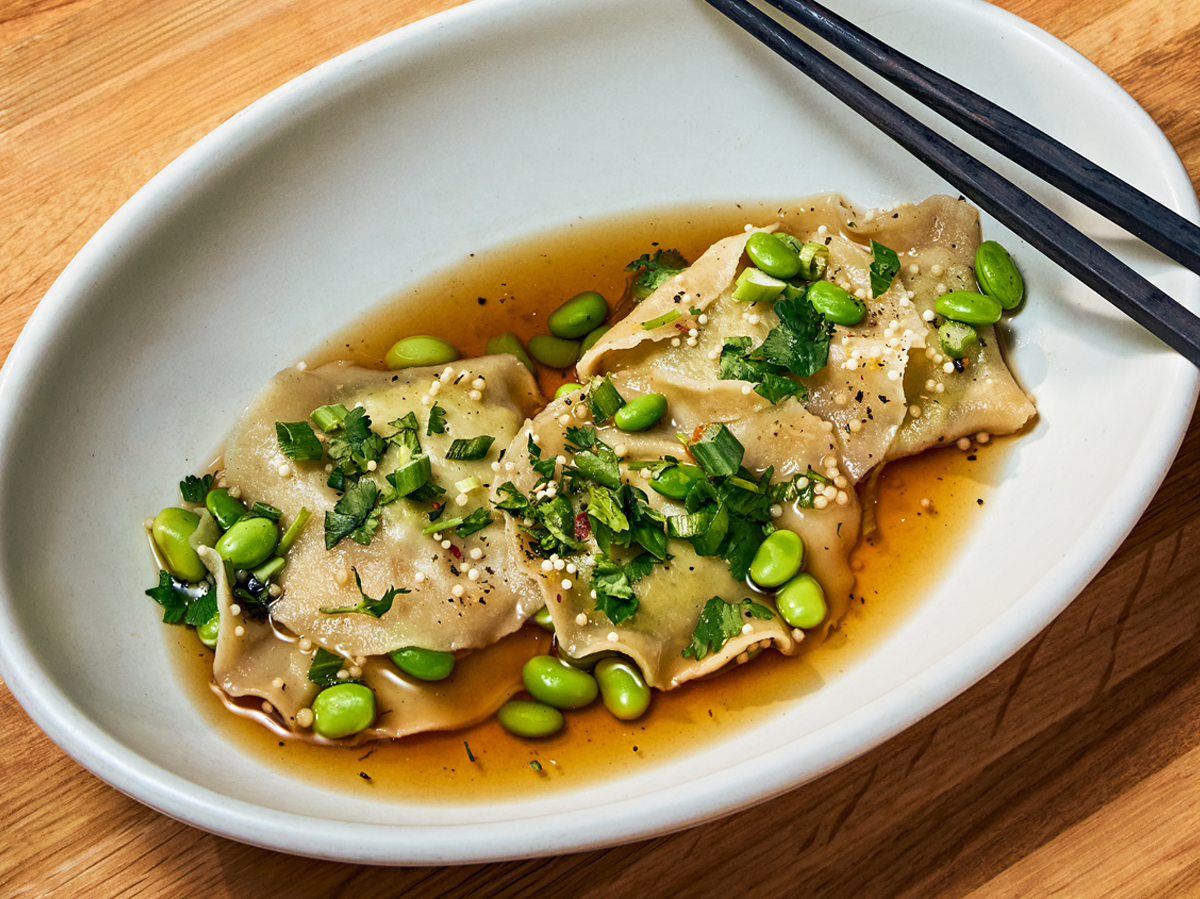 A plate of dumplings garnished with herbs and edamame, served in soy-based sauce, accompanied by chopsticks on a wooden table.