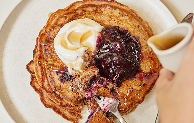 Pancakes topped with yogurt, berry compote, and syrup are served on a plate. A person pours syrup, with a fork cutting a bite.