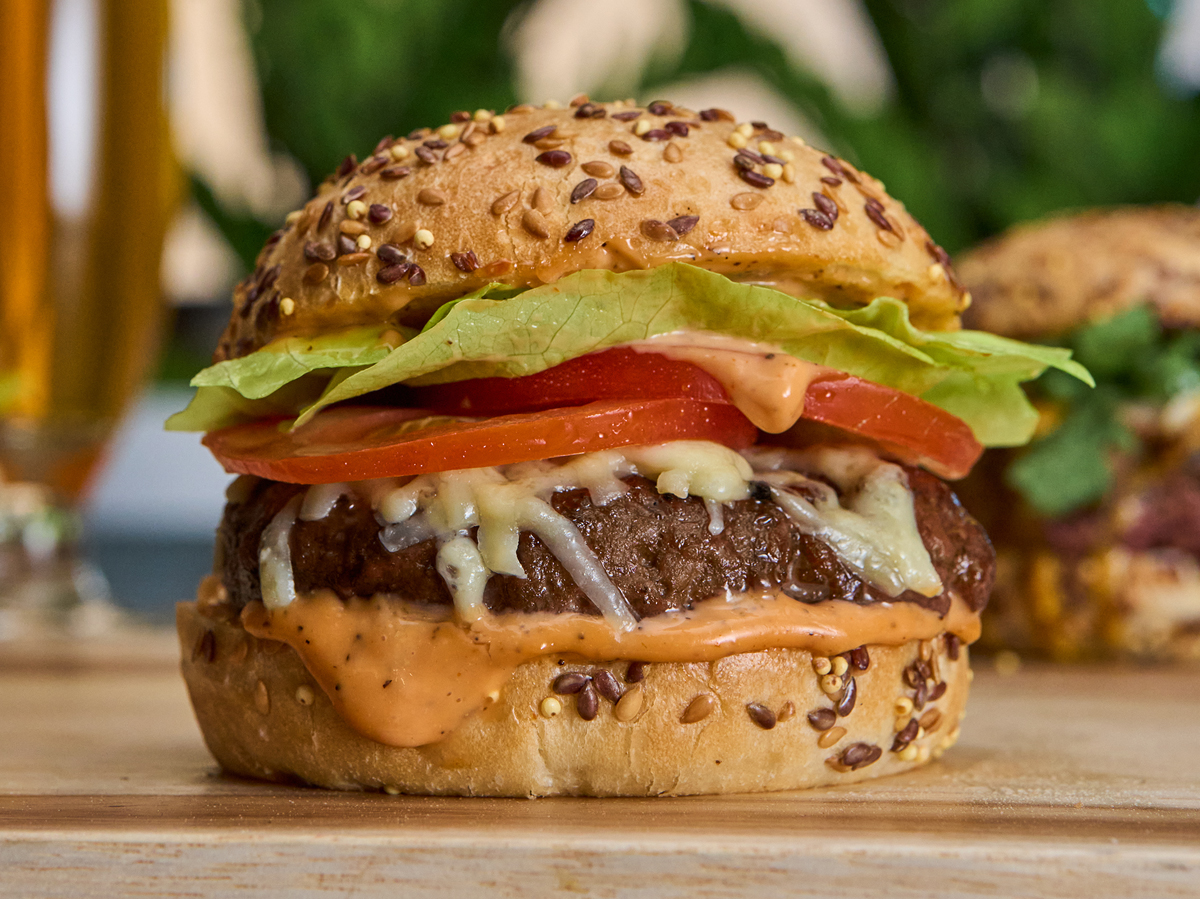A close-up of a freshly made cheeseburger with lettuce, tomato, and sauce on a seeded bun, set on a wooden surface.