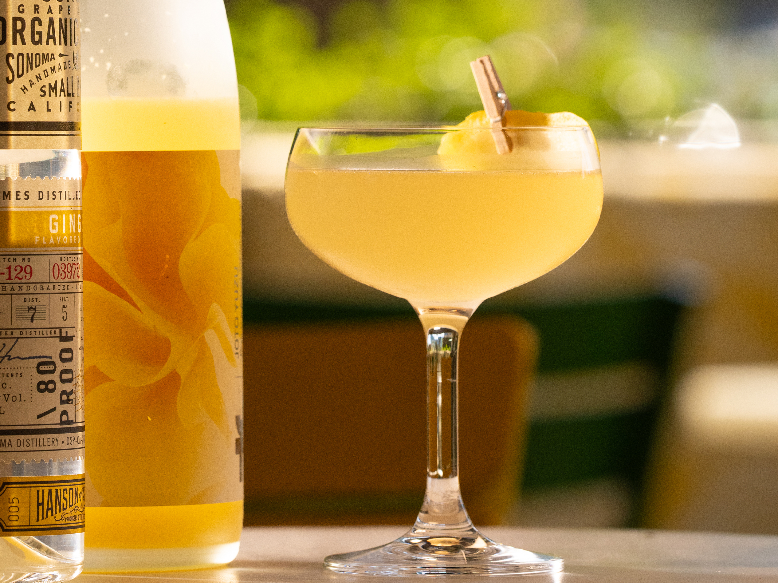 A signature cocktail from True Food Kitchen, featuring a yellow drink in a cocktail glass with a lemon garnish, next to a bottle, set on a table with a bright background."