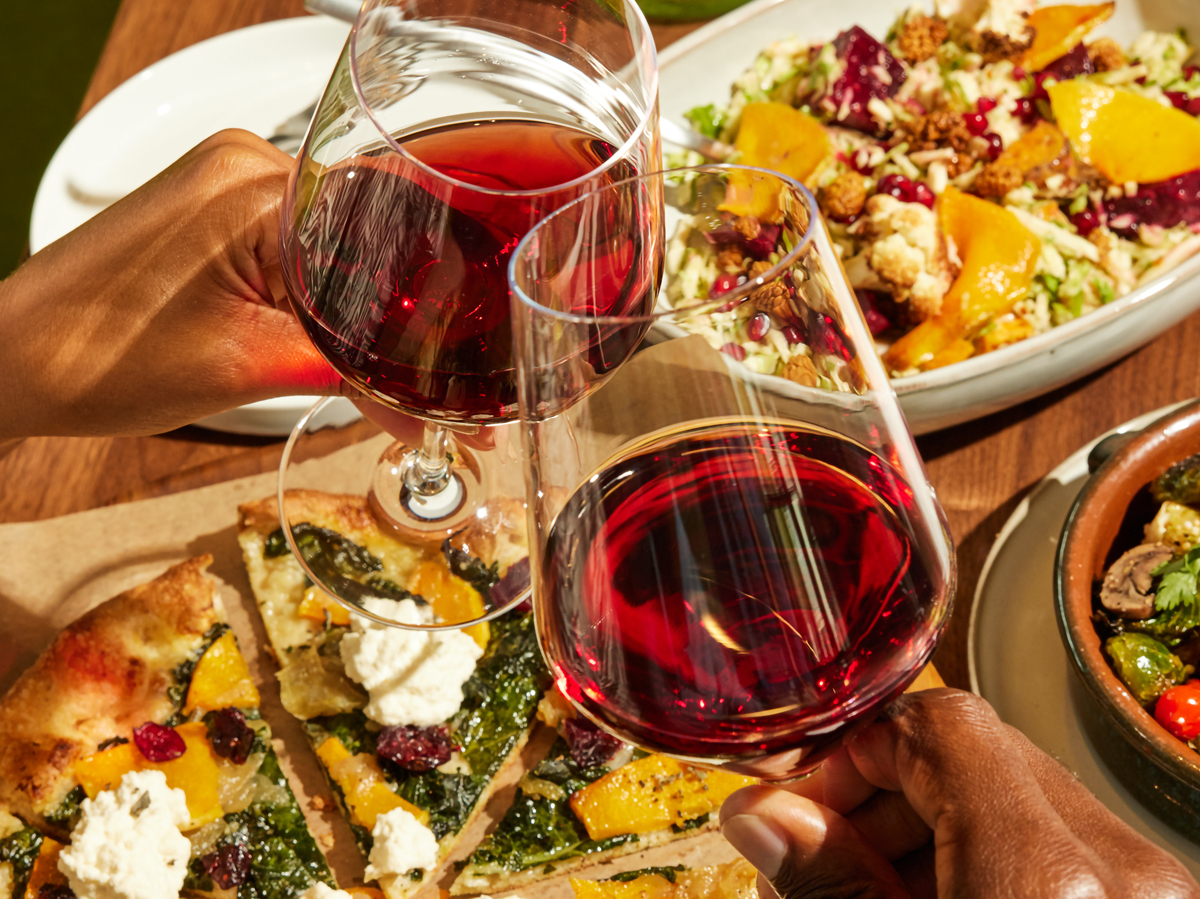 Two people clink glasses of red wine over a table with pizza and salad, creating a warm, inviting dining atmosphere.