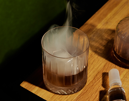 A glass on a wooden table contains a smoking ice cube. Nearby, there's a small dropper bottle, suggesting a scientific or experimental setting.