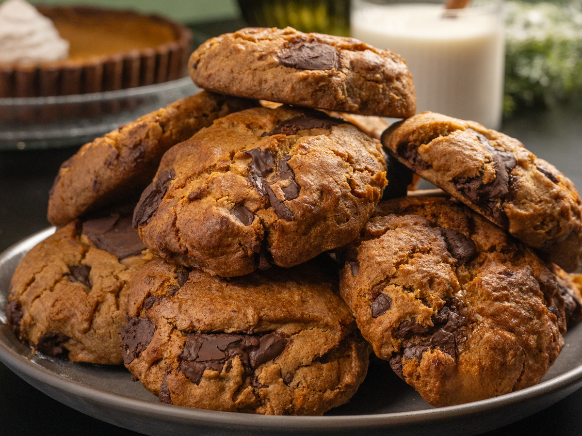 A stack of chunky chocolate chip cookies on a plate, with a glass of milk and a pie in the background.
