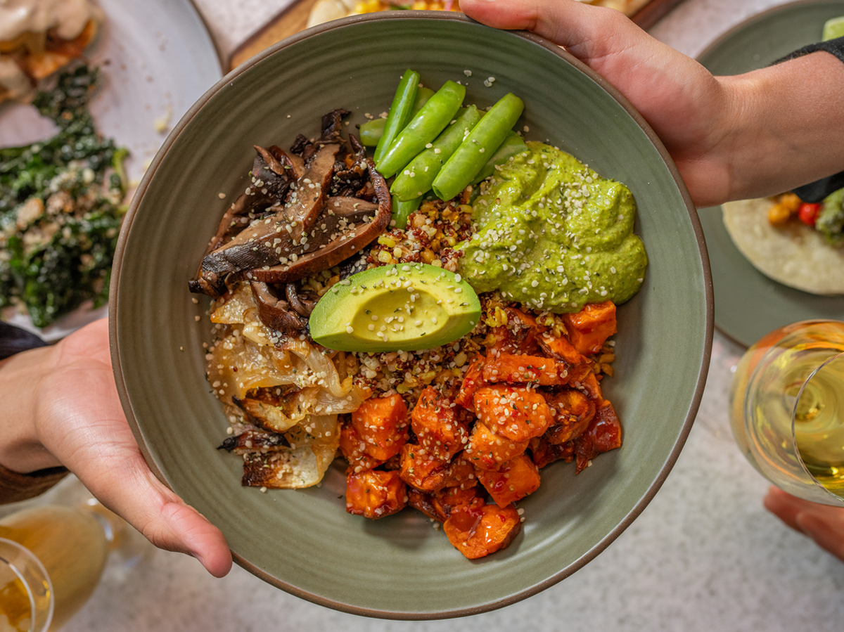 A person holds a bowl of quinoa, avocado, mushrooms, green beans, and sweet potatoes. Another person holds a glass of white wine.