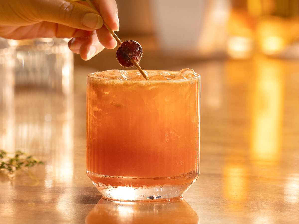 A person holds a cherry-topped cocktail on a bar counter, with warm lighting reflecting off the glass surface.