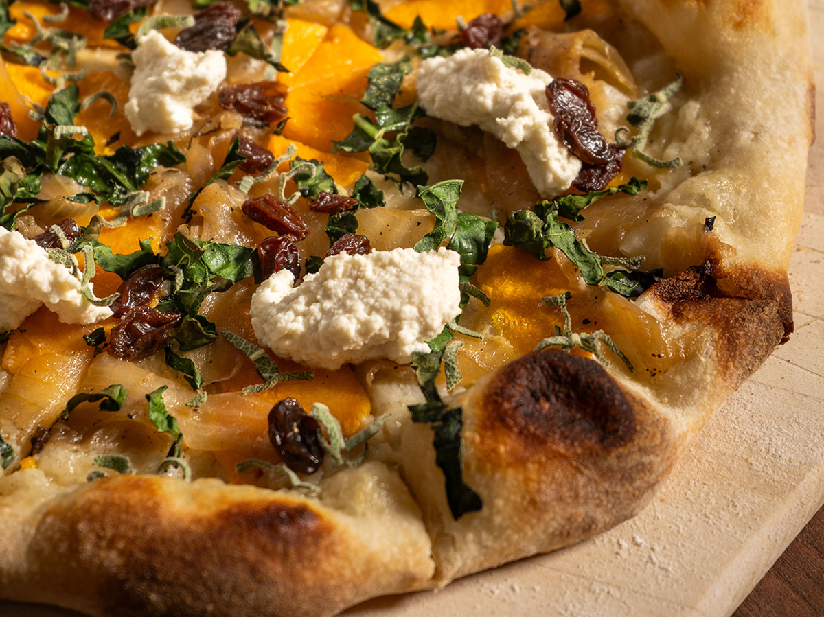 A close-up of a gourmet pizza topped with ricotta, fresh greens, raisins, and a golden crust on a wooden board.