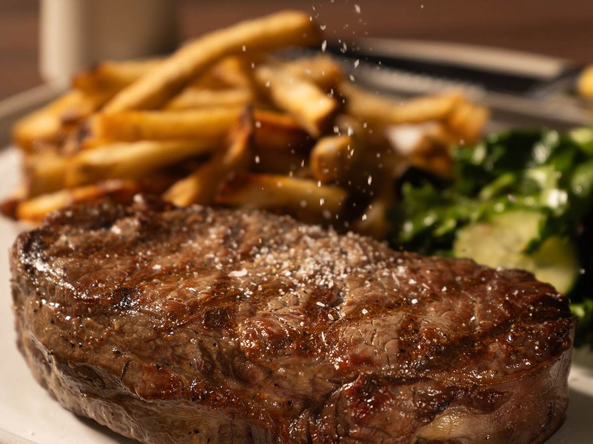 Close-up of a seasoned steak with crispy fries and a fresh salad on a plate. Salt appears to be sprinkled from above.