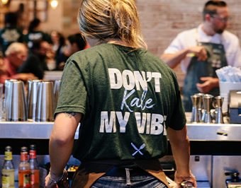 A True Food Kitchen staff member wearing a shirt that says 'Don't Kale My Vibe,' standing behind the bar, with a welcoming and energetic atmosphere in the background.