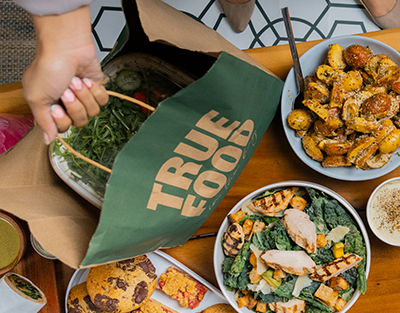 A person carrying a True Food Kitchen catering bag filled with fresh salads and side dishes, showcasing the convenience and variety of the catering options available.
