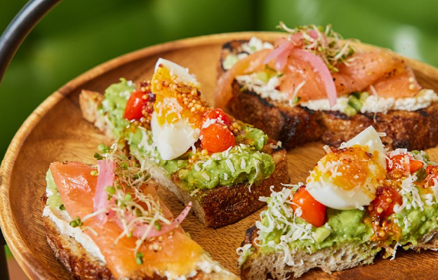 A wooden platter featuring gourmet avocado toasts, topped with salmon, eggs, cherry tomatoes, pickled onions, and sprouts, displayed against a green background.