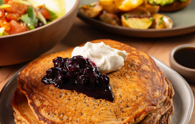 A plate of golden-brown pancakes from True Food Kitchen, topped with a dollop of whipped cream and blueberry compote, served with a cup of coffee and other brunch items in the background.