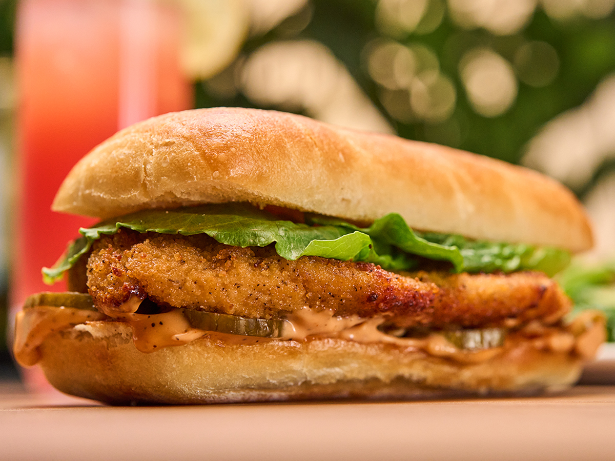 Close-up of a crispy chicken sandwich with lettuce and sauce on a wooden surface; blurred drink and greenery in the background.
