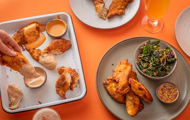 A selection of True Food Kitchen's True Crisp'd Chicken, featuring brined, breaded, and air-fried chicken tenders with avocado oil, served with various dipping sauces and kale slaw, set against an orange background.