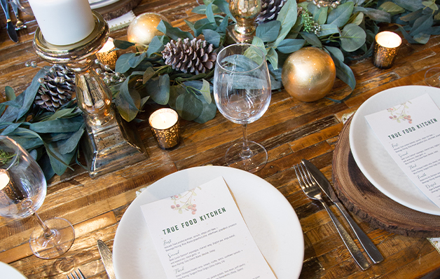 Elegant dining table with pinecone and eucalyptus decor, candles, and menus from "True Food Kitchen" on wooden plates, set for a festive meal.