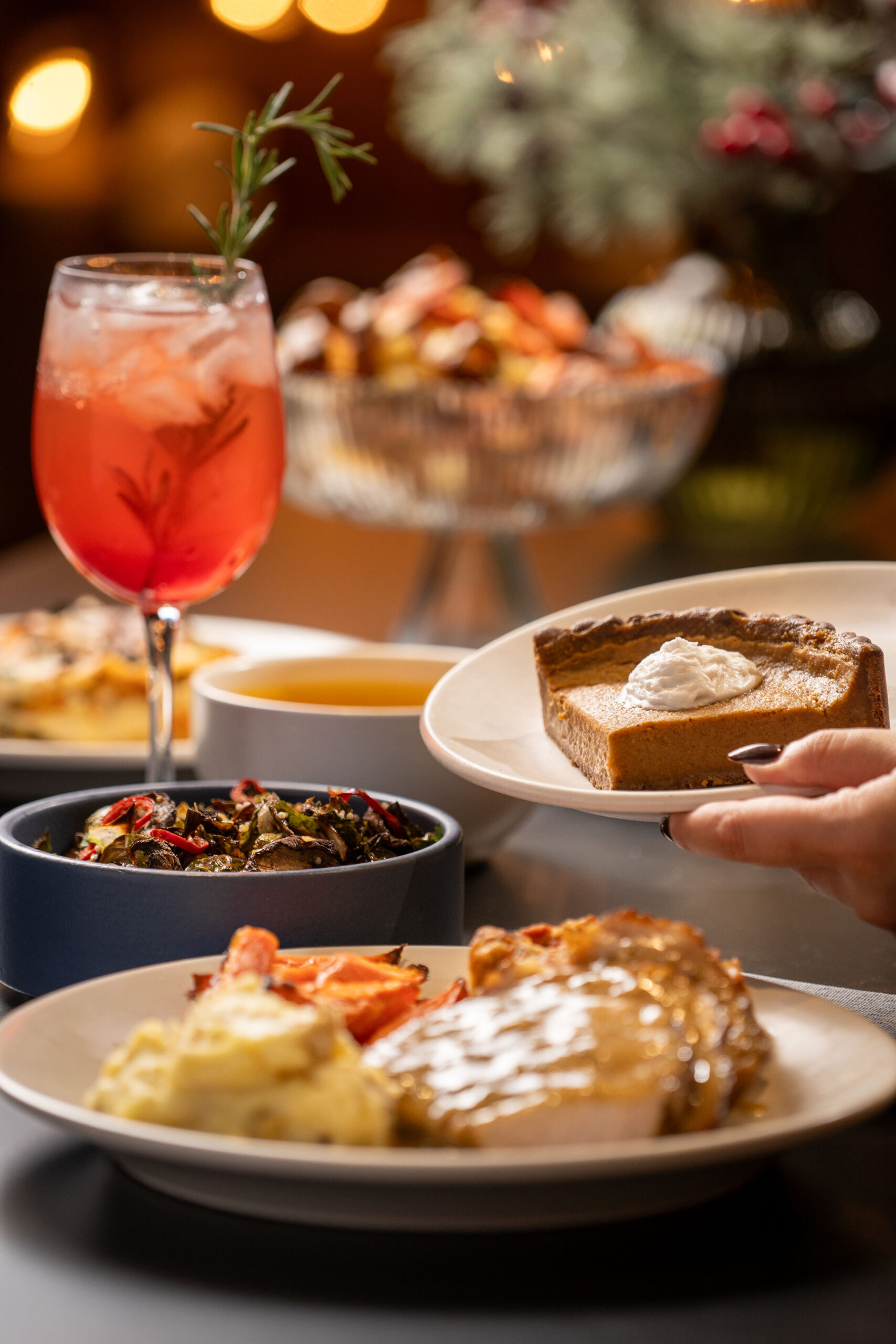 A table filled with assorted dishes, including a slice of pie, colorful vegetables, roast, and a red beverage garnished with rosemary.