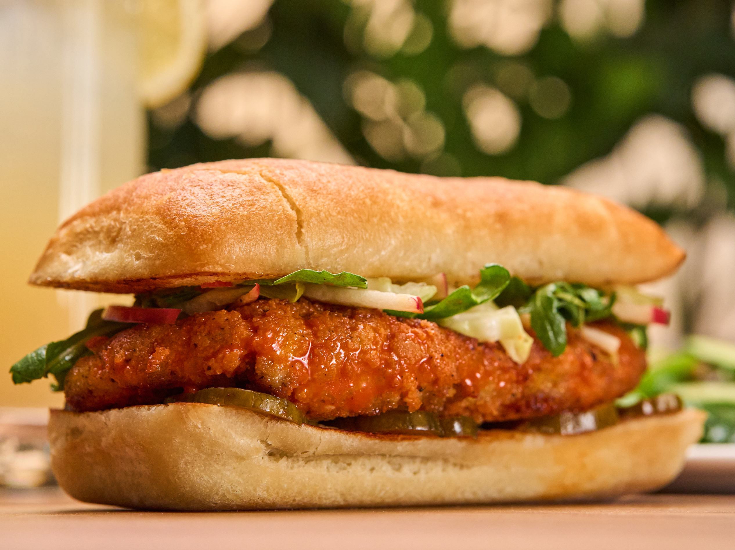 The Spicy Korean Chicken Sandwich from True Food Kitchen, featuring crispy chicken in a brioche bun with house-made pickles, Korean BBQ sauce, and kale slaw, set against an orange background.
