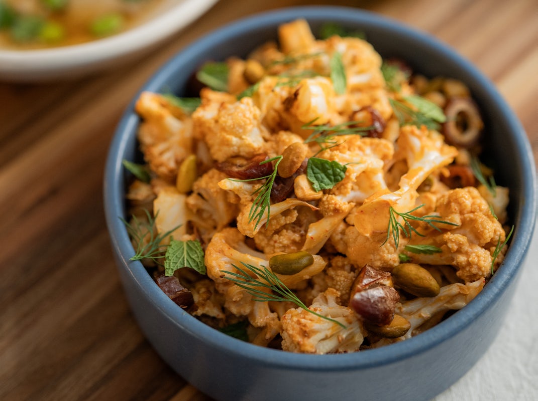A blue bowl filled with roasted cauliflower, olives, herbs, and seeds on a wooden table, alongside a partial view of another dish.