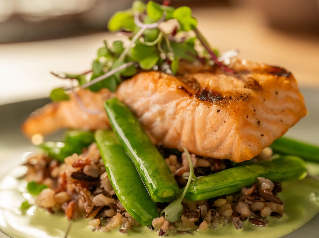 A gourmet dish featuring grilled salmon atop wild rice, snap peas, and a light green sauce, garnished with fresh microgreens.
