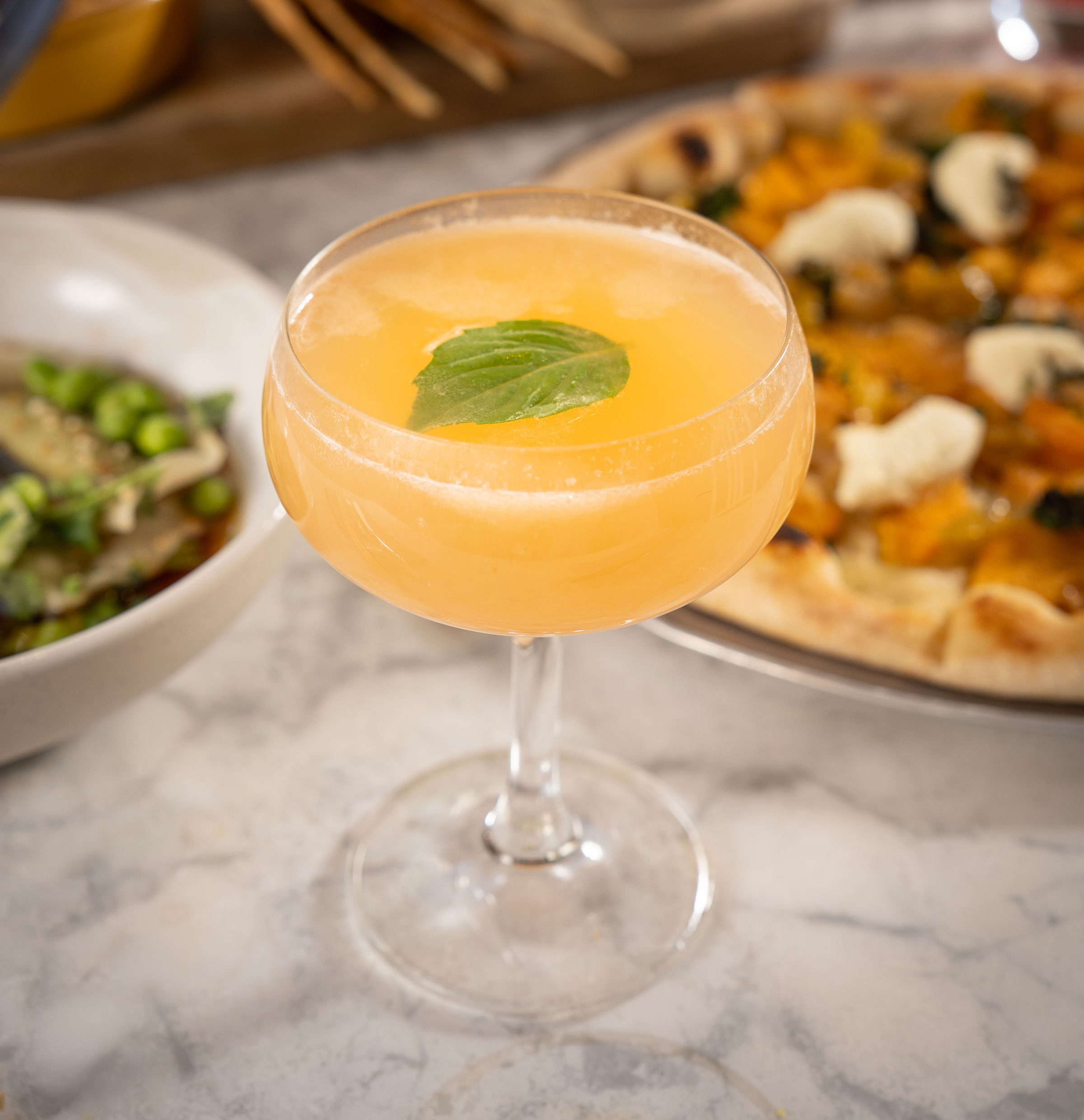 A refreshing orange cocktail garnished with a basil leaf is placed on a marble table, accompanied by pizza and a bowl of greens.
