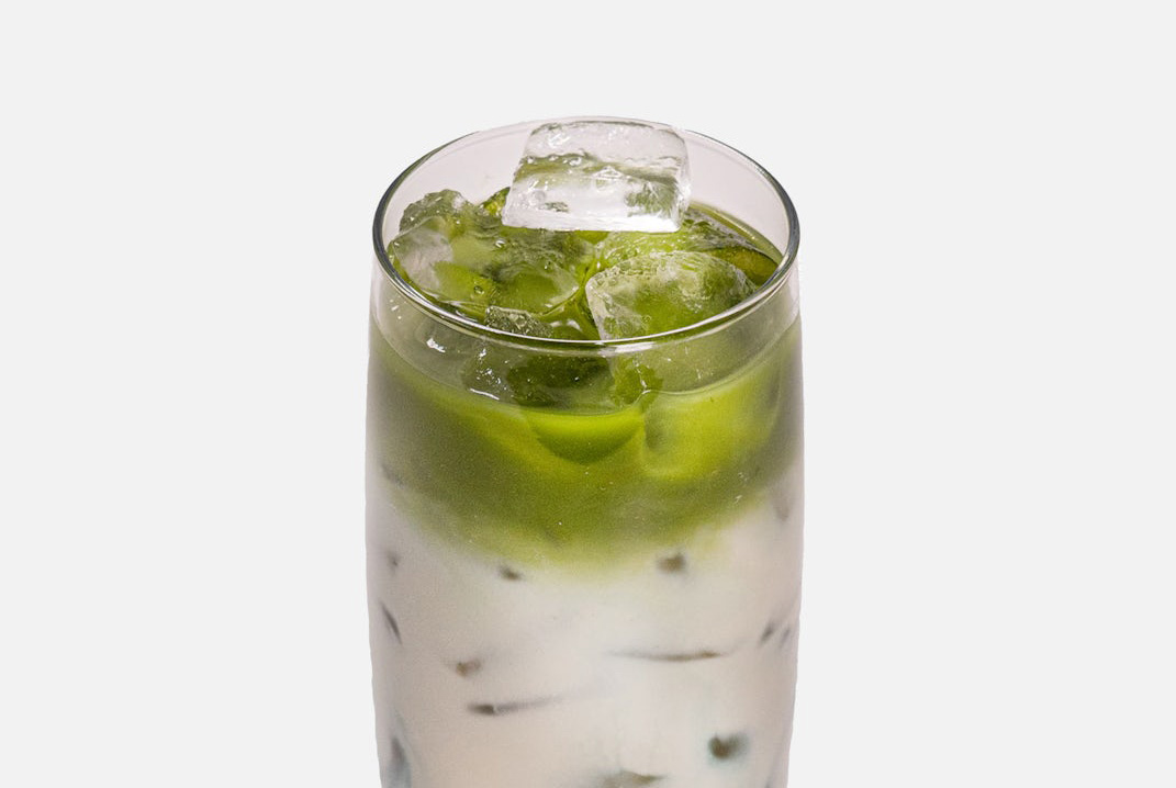 A tall glass filled with a layered iced drink featuring matcha and milk, topped with ice cubes, on a plain background.