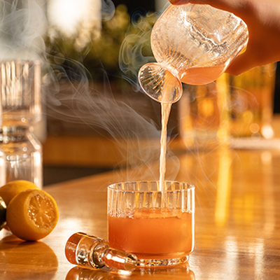 A person pours a steaming beverage into a glass on a wooden bar. Lemons and a small dropper bottle are nearby.