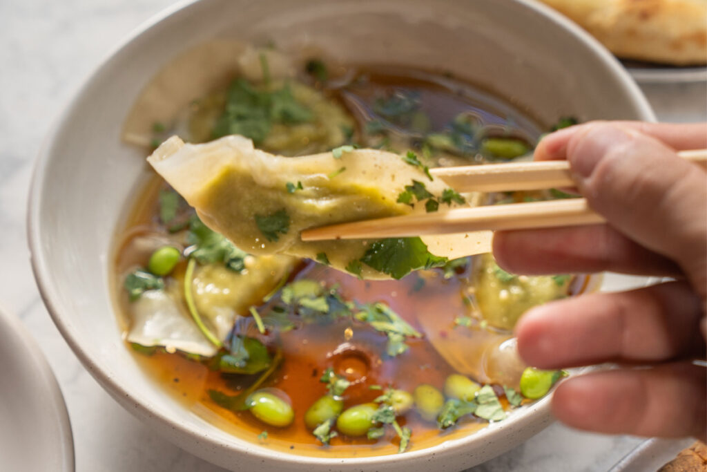 A person holding dumpling with chopsticks over a bowl of broth, garnished with cilantro and edamame, on a light-colored table.
