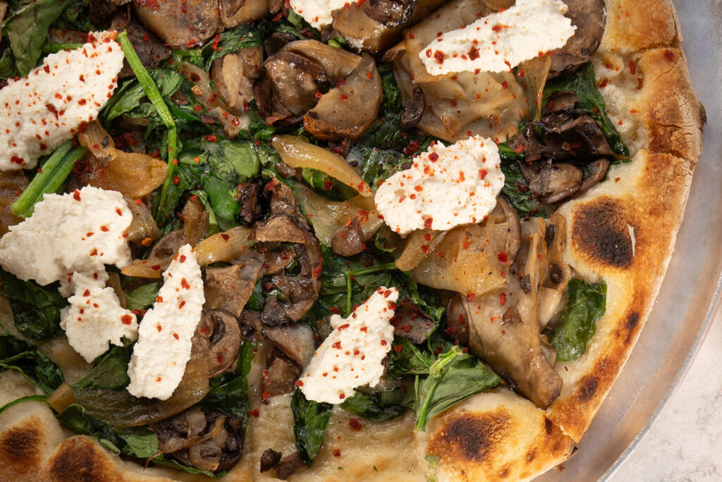 Pizza topped with mushrooms, spinach, onions, and dollops of ricotta cheese on a crispy crust. Sprinkled with red pepper flakes.