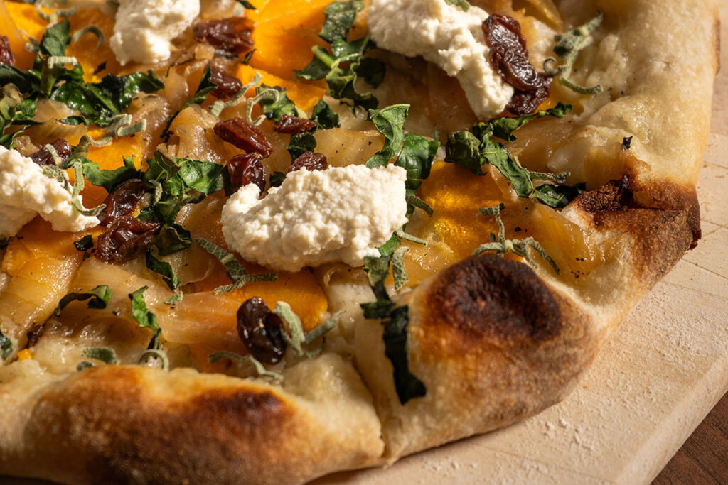 A close-up of a pizza featuring cheese, greens, caramelized onions, and raisins on a wooden board, highlighting its crispy crust and toppings.