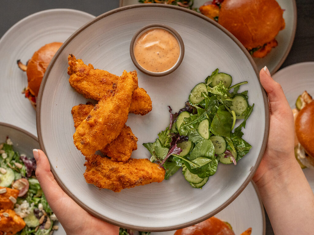 A person holds a plate with crispy chicken strips, green salad, and dipping sauce. Additional plates with burgers are partially visible.