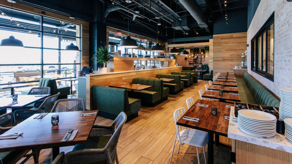 Interior view of a True Food Kitchen restaurant, featuring wooden tables neatly set with rolled napkins, metal chairs, and a green cushioned bench. The restaurant is bright and spacious with large windows, modern decor, and a vibrant mural on the wall that reads 'Eat Better, Live Better.